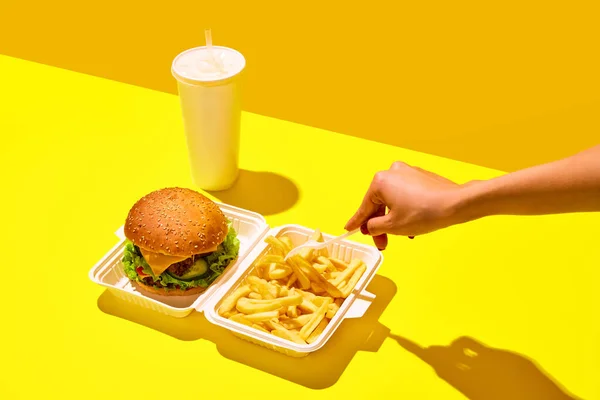 Unpacking tasting fast-food. Unrecognizable woman eat hamburger with soda and fried potato in paper boxes against vivid yellow background. Concept of junk food, menu, delivery, catering, take away. Ad