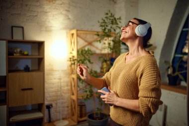 Oturma odasında kulaklıkla müzik dinlerken örgü örüp dans eden muhteşem son sınıf hanımının portresi. Aşk kavramı, emeklilik hayatı, emekliler, rahat, tatiller, teknoloji.