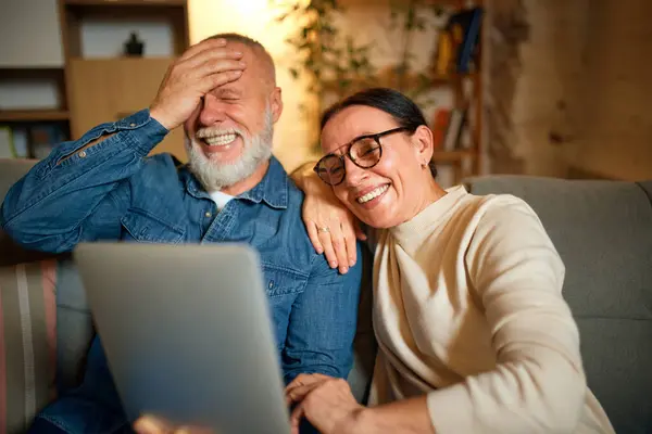 Happy, laughing old lovely couple, elderly man or senior woman watching movies and films on tablet in living room. Concept of love, retirement life, pensioners, cozy, winter holidays, technology