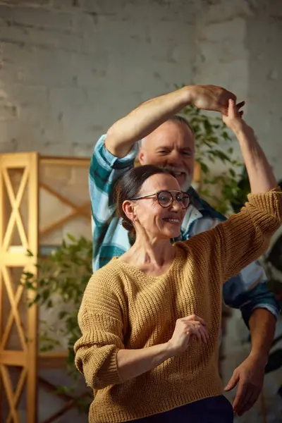 Overjoyed elderly couple dancing in living room at home. Senior wife and husband spending time together. Concept of love, retirement life, pensioners, cozy, winter holidays, relaxing weekend.
