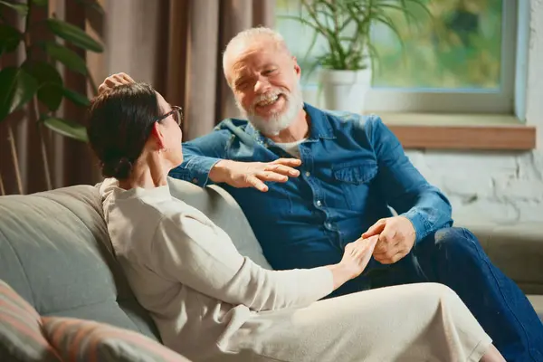 Portrait of happy mature couple in love, wife and husband relaxing while sitting on sofa in living room and talk about life. Concept of love, retirement life, pensioners, cozy, winter holidays.