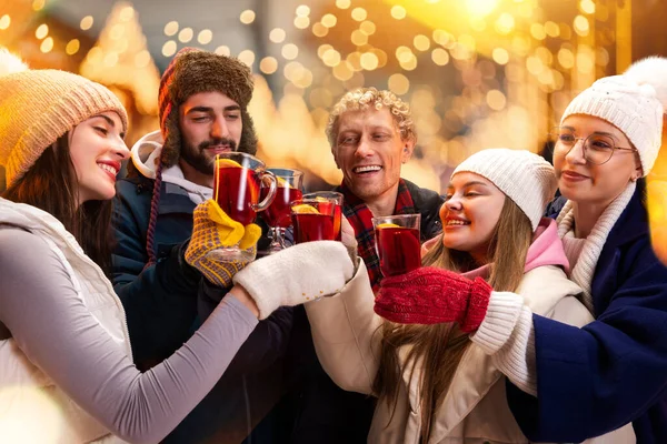 Açık havada duran ve sıcak sıcak bir bardak içeceği tıkırdatan sevimli gülümseyen insanların toplu portresi. Ulusal gelenek kavramı, kış tatili, moda, şenlik