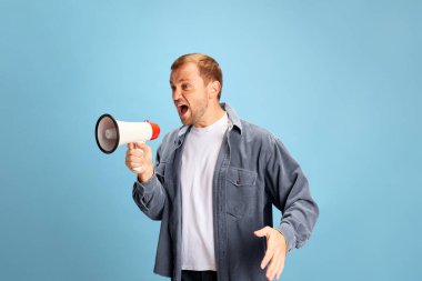 Portrait of angry crazy wild young man shouting to loudspeaker isolated blue studio background. Irritated young guy in casual outfit. Concept of facial expression, human emotions, sale. Copy space, ad clipart