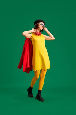 Moda görünümlü, güneş gözlüklü ve kulaklıklı güzel bir kadının portresi parlak yeşil stüdyo arka planında izole edilmiş kırmızı bir ceket taşıyor. Moda anlayışı, tarz, olumlu insani duygular. Boşluğu kopyala