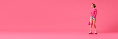 Pankart. Gülümseyen güzel bir kadın, modern aksesuarlarla moda kıyafetleri giyen ve poz veren bir model izole edilmiş pembe stüdyo arka planında paten kayarak eğleniyor. İnsani duygular, tarz, geçmişe dönüş kavramı.