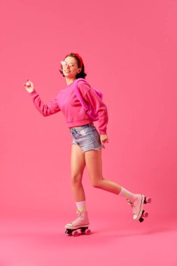 Genç bir kadının tam boy portresi, modern pembe aksesuarlarla moda kıyafeti giyen mankenler izole edilmiş canlı stüdyo arka planında paten kayarken eğleniyorlar. Gençlik kavramı, tarz, insani duygular. ad