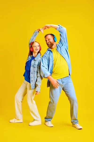 Father and daughter, parent and teen dressed style denim outfit and make hear from arms while listening music in headphones isolated yellow background. Concept of parenthood, childhood, family, style.
