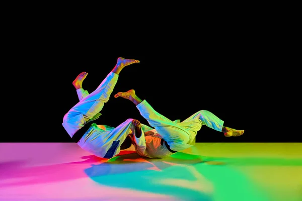 Martial arts match. Professional karate fighters, sportsmen in uniform performing kicks during competition in neon light isolated black background. Concept of martial art, combat sport, energy, fit.
