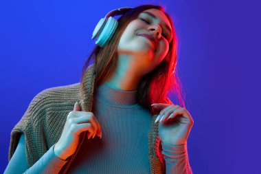 Güzel, aşırı neşeli bir kız, kulaklıkla müzik dinleyen bir öğrenci. Mavi stüdyonun arka planında, neon ışıkta, filtrede. Güzellik anlayışı, insani duygular, yüz ifadesi, tarz. Ad