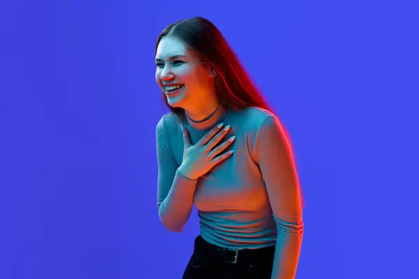 Positive emotions. Young attractive girl, laughing teenager, smiling student isolated blue background in neon filter, light. Concept of beauty, human emotions, facial expression, fashion and style. Ad