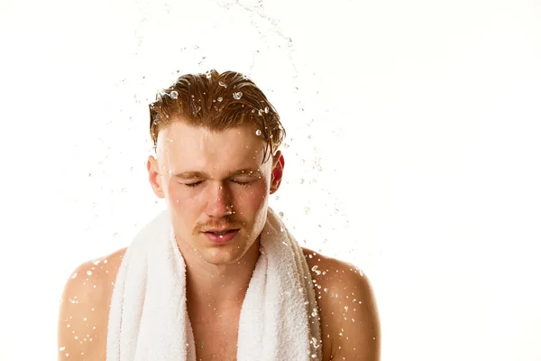 Morning routine. Pleased, handsome red haired male model washing face in splashes of water against white studio background. Concept of natural beauty, selfcare, body and skincare, hygiene, fashion.