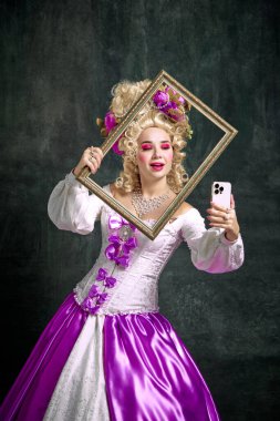 Klasik tarih karakteri gibi giyinmiş, eski moda elbiseler giymiş, elinde retro çerçeve tutan ve klasik arka planda cep telefonuyla selfie çeken bir kadın. Moda kavramı, tarz, modern yaratıcılık.