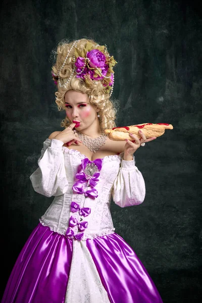 Woman dressed like classical medieval person, queen eating hot, delicious and huge American hot dog against vintage background. Concept of food, nutrition, art, catering, restaurant menu, delivery.