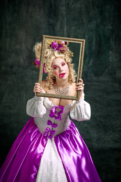 Woman dressed like classic historical character, in old-fashioned dress holding retro frame and grimacing looking at camera over vintage background. Concept of fashion, style, historical, creativity,