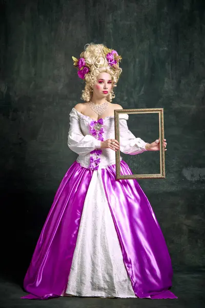 Beautiful woman, aristocratic medieval person in old-fashioned dress holding retro frame over vintage background. Concept of fashion, style, cosplay, historical, creativity, comparisons of eras.
