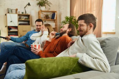 Grup fotoğrafı. Erkekler ve kız korku filmi izliyor, Cadılar Bayramı akşamında korku filmi, evdeki oturma odasında kanepede oturuyor. Arkadaşlık kavramı, birliktelik, online etkinlik, eğlence.