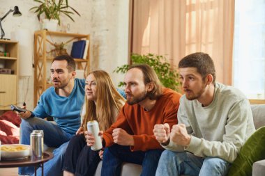 Çekici genç insanlar, erkek ve kadın televizyonun önünde oturuyor ve dikkatle izliyorlar, futbol maçı izliyorlar ve alkollü içeceklerle atıştırmalık yiyorlar. Arkadaşlık kavramı, birliktelik, online etkinlik.