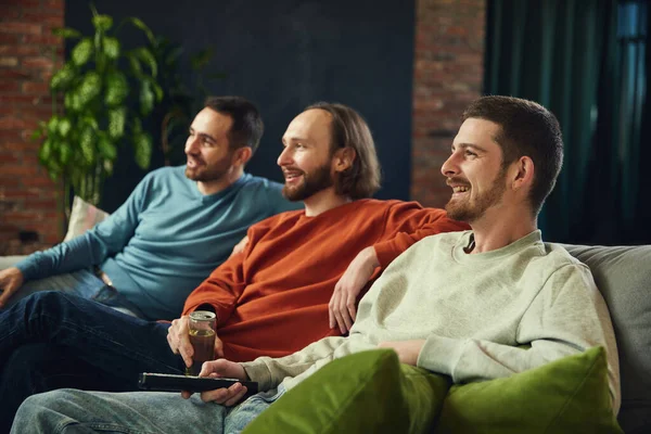 stock image Happy and cheerful young men with remote control and watching football match sitting on coach in living room at home. Concept of friendship, togetherness, TV program, online event, recreation.