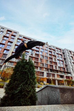 Sporcu, yüksekten atlayan genç bir adam, gökyüzü manzarası üzerinde yüksek binalar arasında parktaki duvarda taklalar atıyor. Göz balığı filtresi. Yaşam tarzı, spor, serbest stil, aktivite, hareket kavramı. Ad