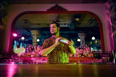 Genç adamın seçici odak noktasında kokteyl karıştırıcı sallarken modern tasarım ve neon aydınlatma ile barda tatlı kokteyller yaparken çekilmiş bir fotoğraf. Parti zamanı, gece kulübü, insanların yaşam tarzı..