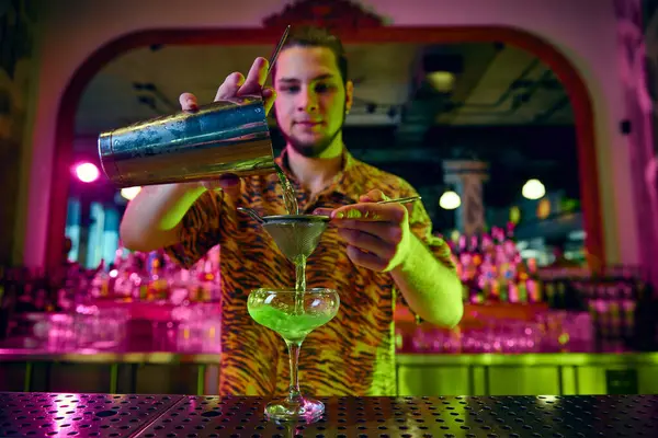 Young guy, bartender made sweet tasty cocktail and pouring it in cocktail glass at bar with modern design and neon illumination. Concept of party time, night club, people lifestyle, work and job.