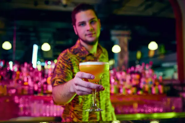 Portrait with selective focus. Young guy, bartender holding, serving sweet tasty cocktail at bar with modern design and neon illumination. Concept of party time, night club, people lifestyle, work.