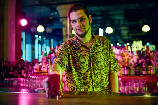 Bartender in fashion stylish shirt serving a bloody mary in cocktail glass on bar counter in nigh club with neon illumination. Concept of party time, night club, people lifestyle, work and job.