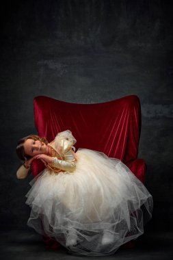 Kırmızı tahtta oturan sevimli küçük kız eski moda elbiseler içinde ortaçağ insanı gibi giyinmiş ve eski moda eskimiş arka planda hayal kuruyor. Tarihi yeniden yapılanma kavramı, dönemlerin karşılaştırılması, duygular.