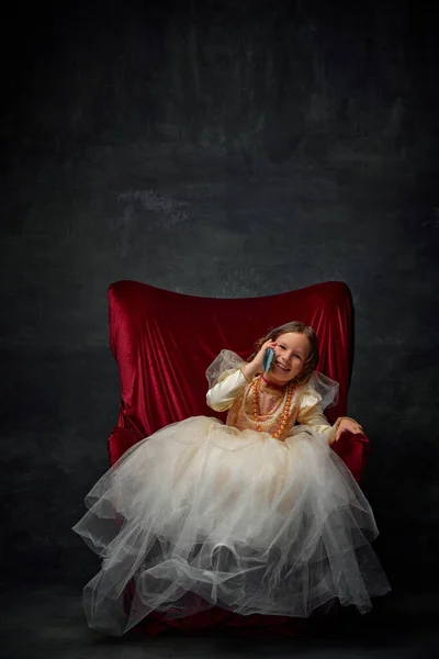 Little blondie girl, in costume of royal person lying in red throne and talking on phone and laughing against dark vintage background. Concept of historical, comparison of eras, medieval fashion.