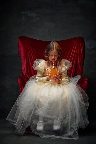 capricious queen. Upset little girl, dressed in festive costume of royal person and dont want to drink soda against dark vintage background. Concept of comparison of eras, medieval fashion, emotions.