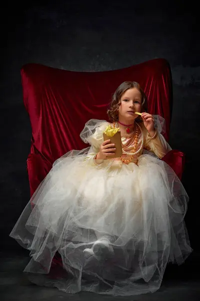 Little princess eating, tasting hot fries potato at fist time sitting in royal throne against dark vintage background. Concept of historical remake, comparison of eras, medieval fashion, emotions.