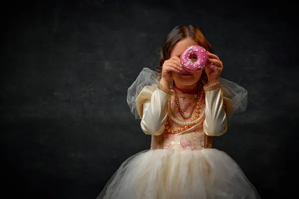 One little medieval person, small girl dressed in old-fashioned costume peeking through donut hole against dark vintage background. Concept of historical remake, comparison of eras, medieval fashion.