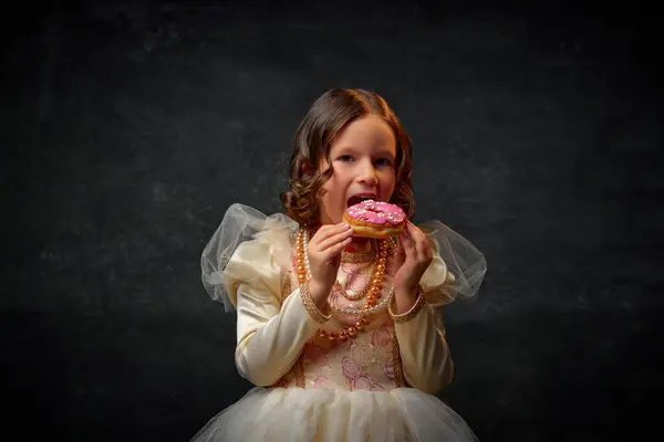 One little medieval person, small girl dressed in old-fashioned costume eating sweet tasty donut against dark vintage background. Concept of historical remake, comparison of eras, medieval fashion.