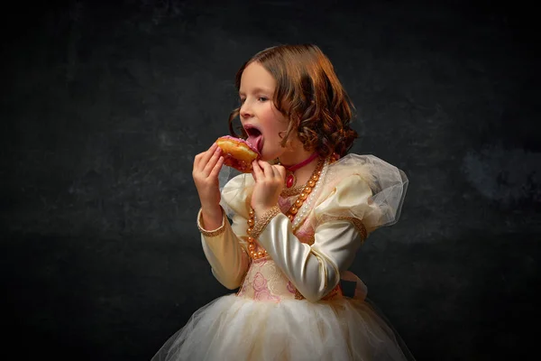 One little medieval person, small girl dressed in old-fashioned costume licks sweet pink icing from donut against dark vintage background. Concept of remake, comparison of eras, medieval fashion.