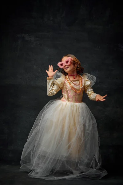 Full-length portrait of royal person, small girl dressed in old-fashioned costume with modern accessories an dancing against vintage background. Concept of comparison of eras, medieval fashion.