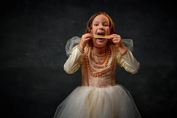 Portrait of little medieval person, small girl dressed in old-fashioned costume bites sweet tasty candy against vintage background. Concept of historical remake, comparison of eras, medieval fashion.