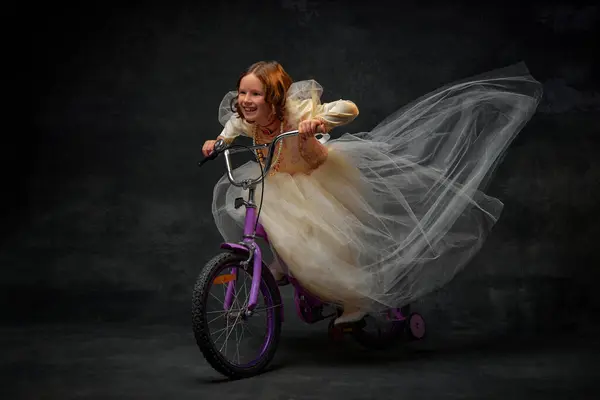 Charming little princess, dressed like medieval person riding childrens bicycle and having fun against dark vintage background. Concept of historical remake, comparison of eras, medieval fashion.