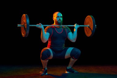 Kaslı, güçlü genç bir adam, vücut geliştirici, çömelme ve halter kaldırma, siyah arka planda ağır yükler. Spor anlayışı, güç, spor, sağlıklı yaşam tarzı, güç, halter