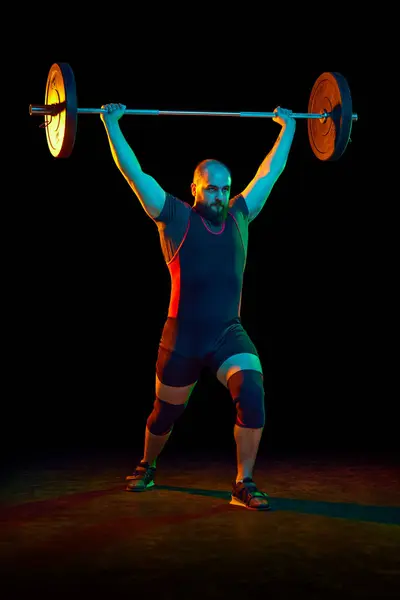 Bearded strong man, athlete training, lifting heavy weights, barbell against black background in neon. Muscular strong body. Concept of sport, strength, gym, healthy lifestyle, power, weightlifting