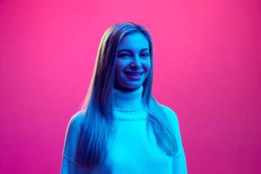 Neon ışıkta pembe arka planda örülmüş kalın süveter giyen, gülümseyen genç ve çekici bir kadının yakın portresi. Güzellik kavramı, insani duygular, yüz ifadesi, moda ve tarz.