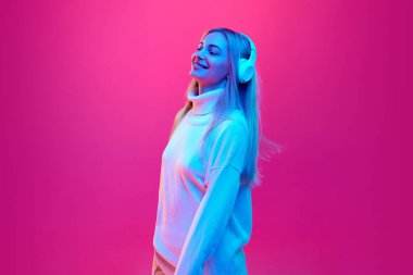 Çekici, neşeli bir kadın, kulaklıkla müzik dinlerken neon ışıkta pembe stüdyo arka planında, filtrede dans ediyor. İnsan duyguları kavramı, enerji, eğlence ve neşe, moda, tarz.