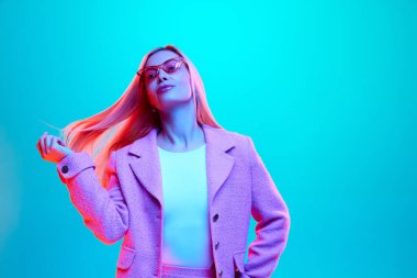 Güzel, şık giyinmiş, pembe gözlüklü genç bir kadının portresi. Neon ışıklı, filtreli, mavi renkli stüdyo arkaplanına poz veriyor. Güzellik kavramı, moda, tarz, iş.