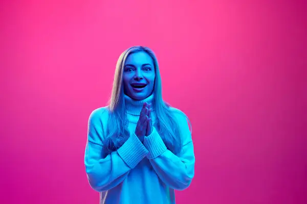 Wow. Surprised, delightful young woman in knitted warm sweater clapping of joy against pink background in neon light. Concept of fashion, style, sales season, winter holidays, fun and joy.