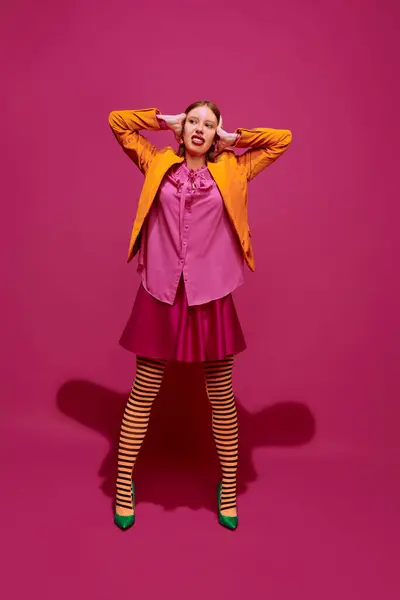Full-length portrait of young attractive girl in unusual, strange, variegated colored clothes and posing against magenta color studio background. Concept of high fashion, style and glamour, beauty, ad