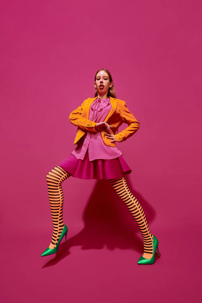 Full-length portrait of young attractive girl in unusual, strange, retro colored outfit and posing against magenta color studio background. Concept of surrealism, art, high fashion, beauty, ad.