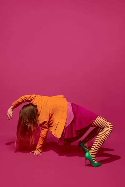 Self-expression. Young weird woman in bright clothes posing in extraordinary pose against magenta studio background. Concept of youth, surrealism, art, high fashion, retro, beauty, ad.
