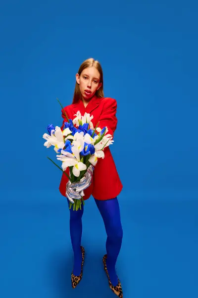 Full-length portrait of young weird girl in unusual, strange, variegated retro clothes and posing with flower bunch against color studio background. Concept of high fashion, style and glamour, beauty.