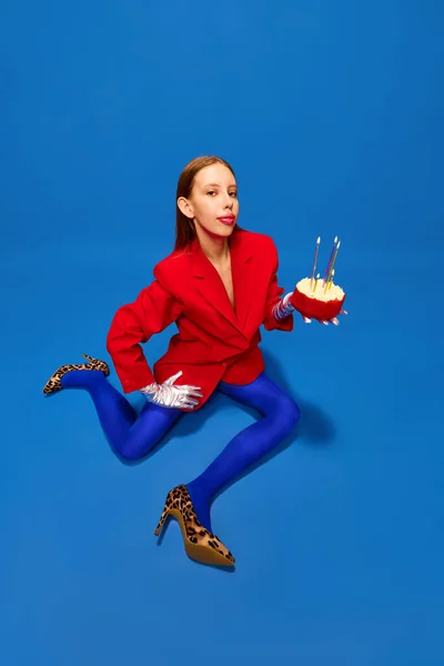 Young lady dressed unusual, freaky, bright outfit, wearing blue tights, red jacket and animal printed heels holding birthday cake with candles. Concept of high fashion, style and glamour, beauty, ad
