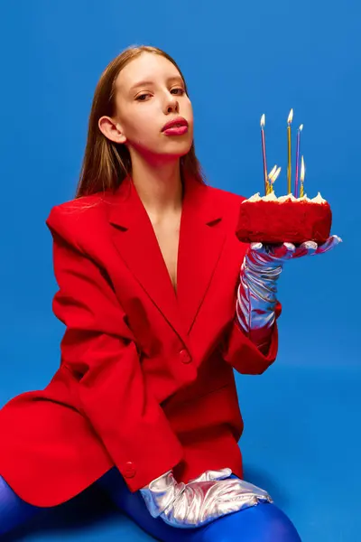 Portrait of young lady dressed unusual, freaky, bright clothes, wearing blue tights, red jacket holding birthday cake looking at camera. Concept of high fashion, style and glamour, beauty, ad