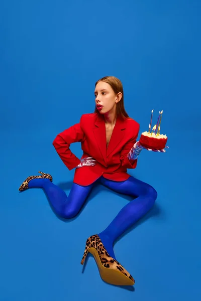 Fashionable verdict. Young lady dressed unusual, freaky, bright outfit, wearing blue tights, red jacket and animal printed heels holding birthday cake. Concept of high fashion, style, glamour, beauty